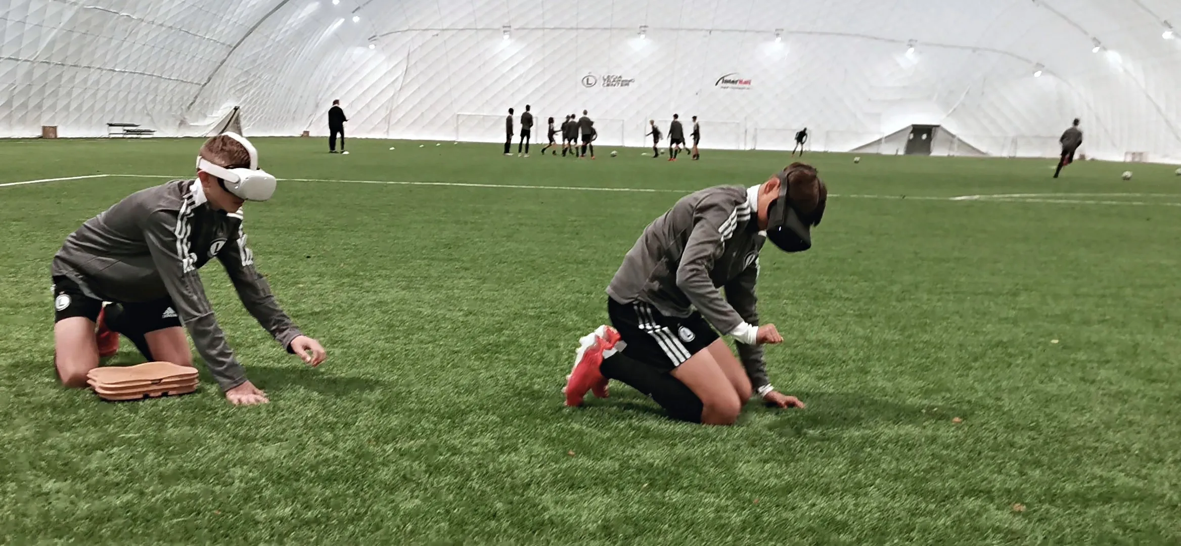 Two young football players in VR headsets kneeling on an indoor pitch and practicing chest compressions