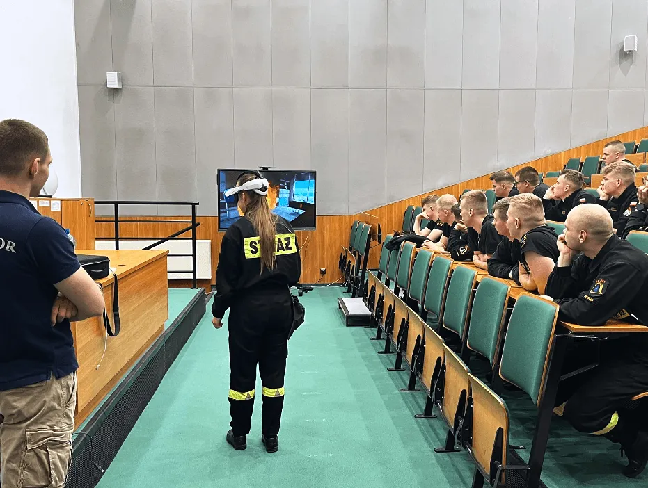A firefighter cadet in a VR headset in front of a lecture hall full of firefighters, with the VR view on a screen
