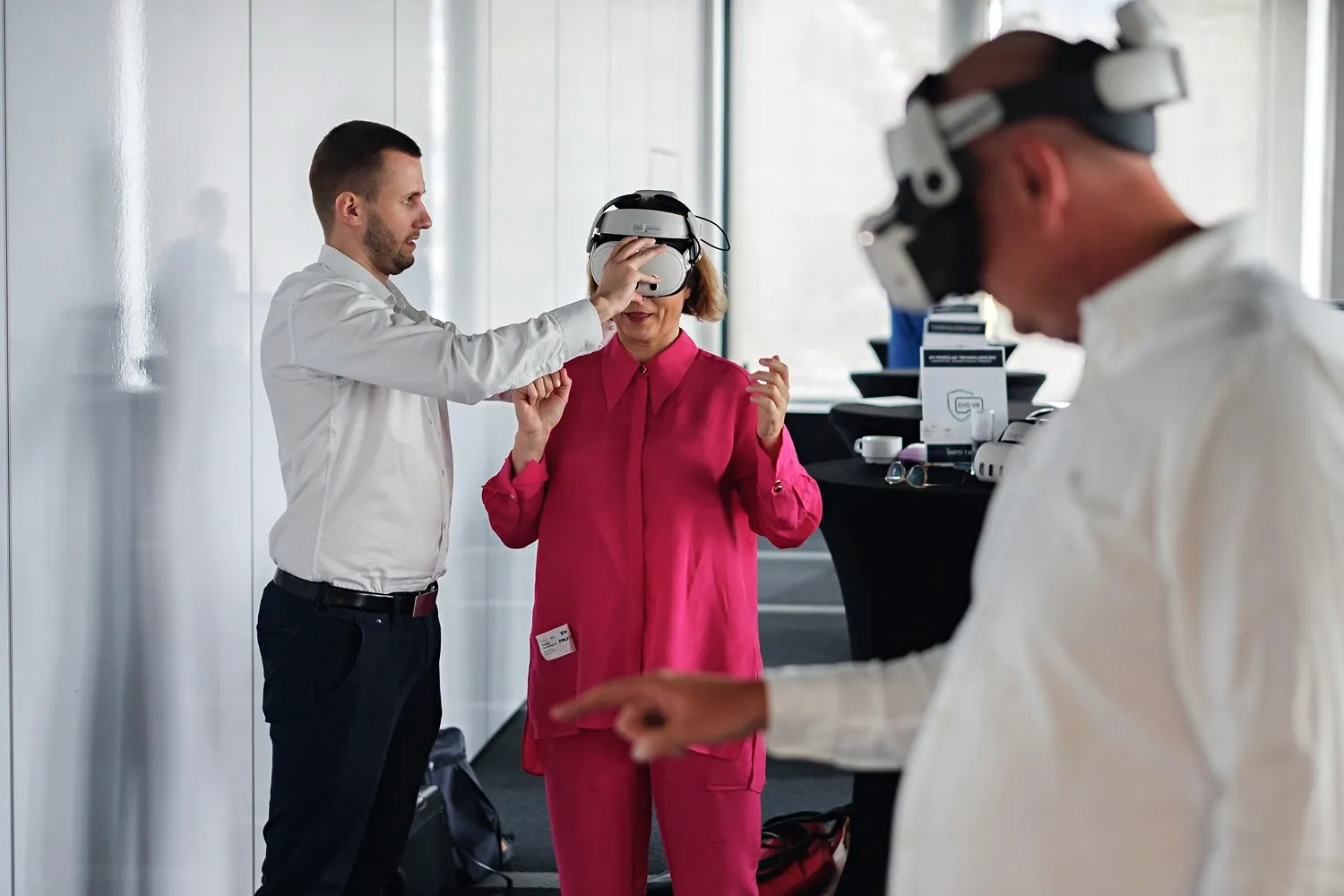 Eine Frau in rosa Anzug und ein Mann in weißem Hemd probieren VR-Brillen aus, während ein Trainer sie anleitet