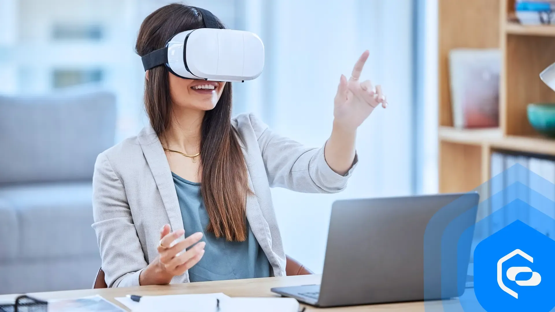 Una mujer sonriente con gafas VR extiende la mano mientras está sentada ante un escritorio con un portátil
